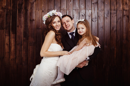 Beautiful pregnant woman in wedding dress and her husband are holding pretty cheerful daughter in arms at a plank wall background. Concept of happy family.の写真素材