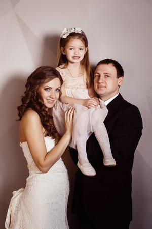 Beautiful pregnant woman in wedding dress and her husband are holding pretty cheerful daughter in arms at a wall background. Concept of happy family.の写真素材
