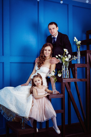 Cute little girl and cheerful father who is tenderly holding his beautiful pregnant wife in wedding dress are sitting on a ladder a blue wall background.の写真素材