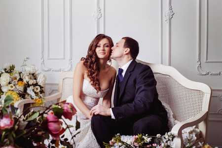 A beautiful wedding pair is kissing and tenderly holding by hands sitting on sofa at home background.の写真素材