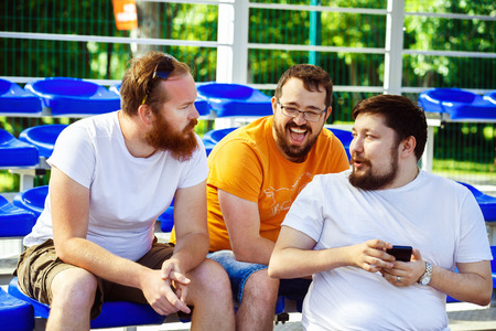 Three male friends are watching mobile phone video and laughing sitting at summer day at stadium background.の写真素材