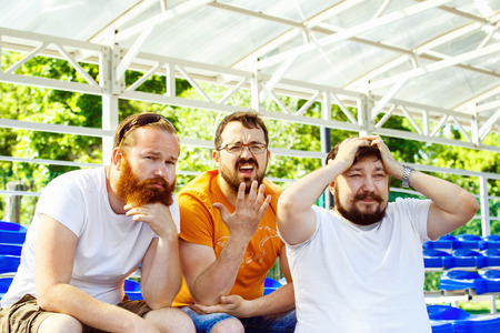 Three bearded football fan friends are sitting upset at goal moment at summer stadium  background.の写真素材