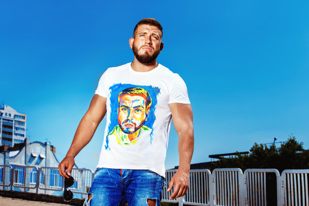 Cool macho bearded man model in jeans and shirt is standing at urban bright summer background.の写真素材