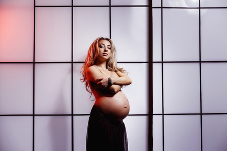 Beautiful pregnant woman is standing indoors at sunset window background.の写真素材