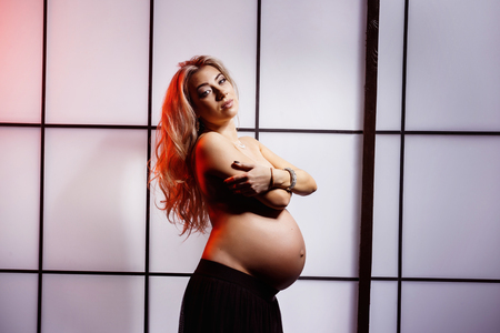 Beautiful pregnant woman is standing indoors at evening window background.の写真素材