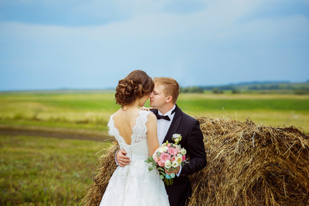 Beautiful young bride is kissing husband at rural haystacks summer field background.の写真素材