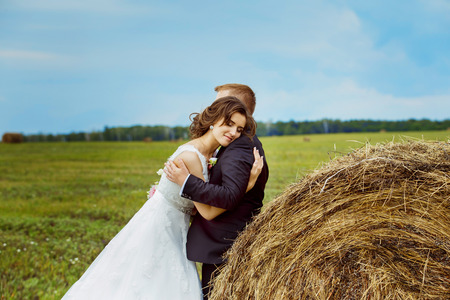 Beautiful young bride with closed eyes is tenderly embracing husband at rural haystacks summer field background.の写真素材
