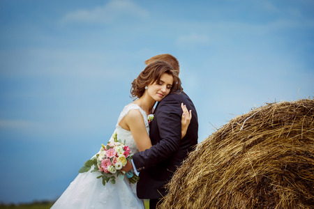 Beautiful young bride with closed eyes is tenderly embracing husband at rural haystacks summer field background.の写真素材