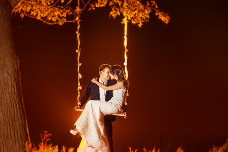 Beautiful young wedding couple is kissing on swing at red sunset background.の写真素材
