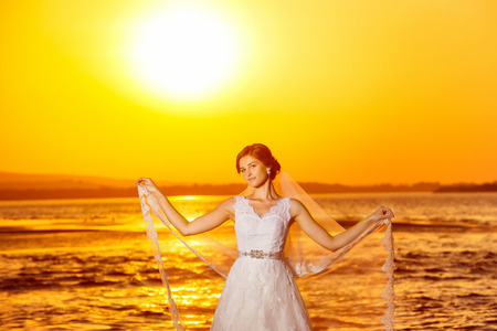 Beautiful bride in long white wedding dress is holding veil at amazing golden sea sunrise background.の写真素材