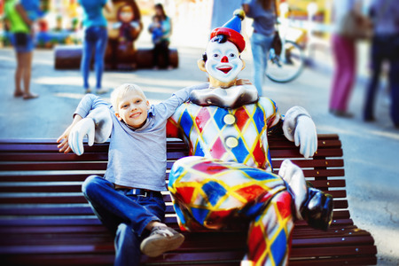 Happy boy is sitting on bench near clown figure at summer holiday park background.の写真素材