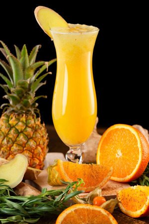 Closeup glass of fresh orange and pineapple juice decorated with ginger and fruits on a wooden table isolated at black background.の写真素材