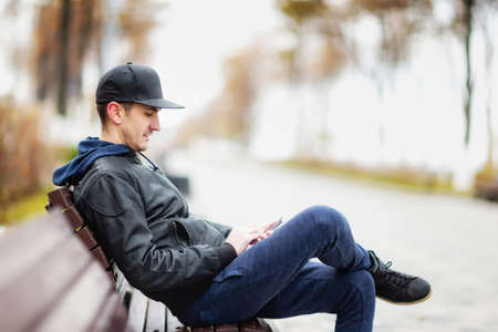 Young man is sitting at a bench with a cellphone in hands and texting outdoors at city park background.の写真素材