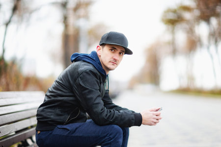 Young man is sitting at a bench with a cellphone in hands and looking at camera outdoors at city...