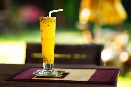 Closeup glass of passion fruit shake served with a straw at a wooden table at tropical outdoors background.の写真素材