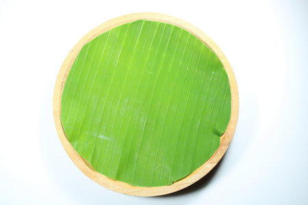 Top View of Round Wooden Plate Lined with Fresh Green Banana Leaf on White Background. Traditional Asian Dining Mockup, Food Presentation, and Minimalist Tropical Culinary Layoutの写真素材