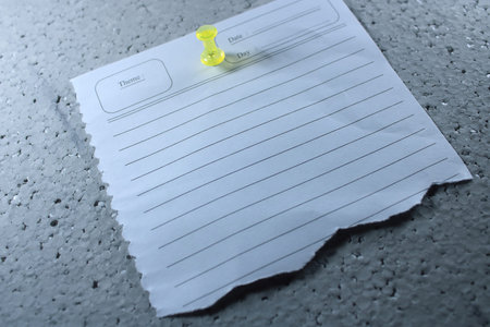 Torn Lined Note Paper Pinned with Yellow Thumbtack on Gray Textured Surface. Minimalist Memo Sheet with Date and Theme Fields for Reminders, Planning, and Message Conceptsの写真素材