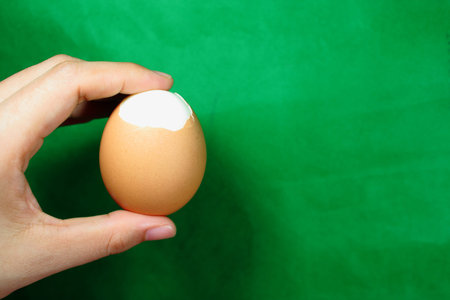 Hand holding a half peeled boiled egg on a green background. Close up of healthy breakfast protein and nutritious eating concept for fitness and culinary themesの写真素材