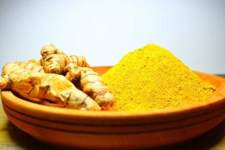 Fresh turmeric roots and ground turmeric powder on a wooden plate, showcasing natural spice used in...