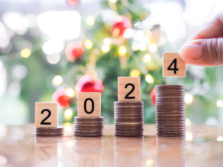 Close up hand putting number 2024 on stack of coins with Christmas light bokeh background. Merry...