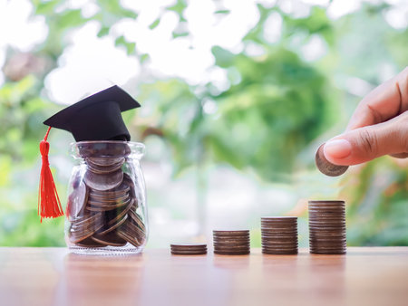 Close up hand putting coin in stack of coins and Glass bottle with graduation hat. The concept of saving money for education, student loan, scholarship, tuition fees in futureの写真素材