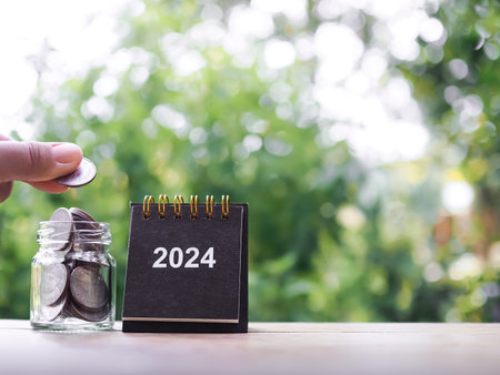 Close up hand putting coin into glass bottle, 2024 desk calendar on the table with copy space. The concept of save money for prepare in new year 2024.の写真素材