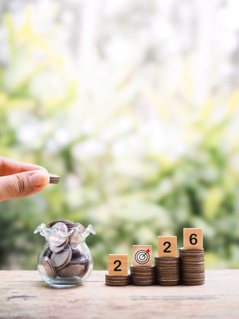 Hand putting coin into glass bottle, Wooden blocks with the number 2026 on stack of coins. The...