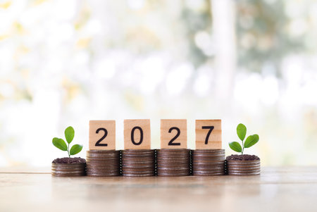 Wooden blocks with the number 2027 and plants growing up on stack of coins. The concept of saving money, Financial, Investment and Business growing in year 2027.の写真素材