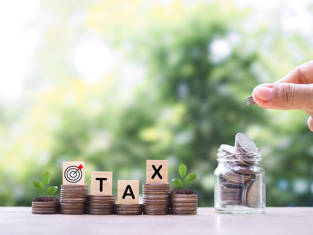 Hand putting coin into glass bottle, Wooden blocks with the word TAX and plants growing up on stack of coins. The concept about saving money and manage time to paying taxの写真素材