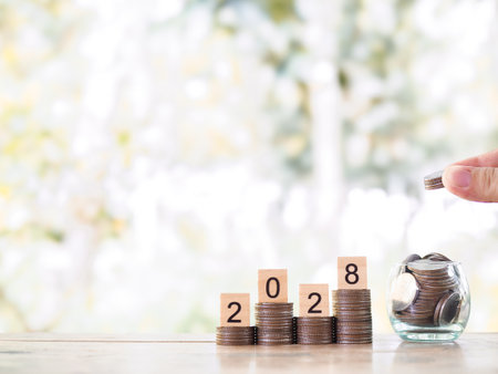 Hand putting coin into glass bottle, Wooden blocks with the number 2028 on stack of coins. The concept of saving money, Financial, Investment and Business growing in year 2028.の写真素材