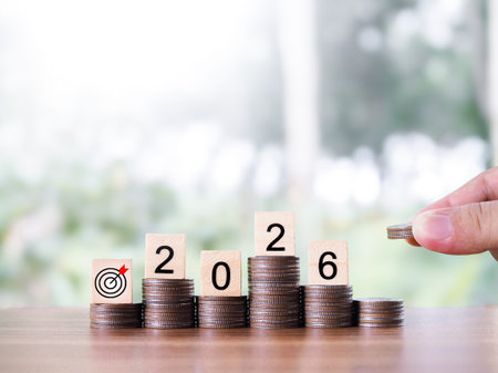 Hand putting coin into stack of coins and wooden blocks with the number 2026. The concept of saving...