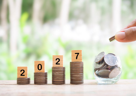 Close up hand putting coin into glass bottle, Wooden blocks with the number 2027 on stack of coins....