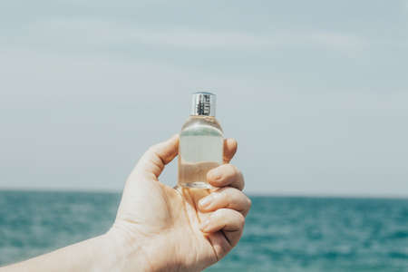 Girls hand with beauty product bottle in front of blue sea on a sunny day. Fresh airy background.の写真素材