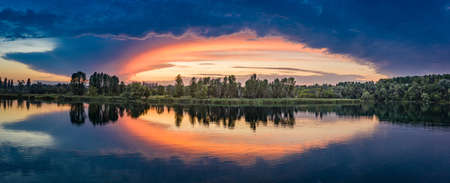 Bright orange-blue sky reflected in the calm clear water of the lake. Fresh summer morning. Picturesque landscape in the countryside.の写真素材