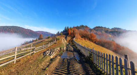 Dirt mountain road in poor condition, with puddles, fenced with an old wooden fence.の写真素材