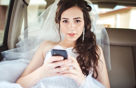 Wedding: beautiful bride sitting in car straight with mobile phone hairstyle and bright makeup. Woman in white dress at wedding day waiting for groom and posing. Concept of love and interest. Newlywedの写真素材