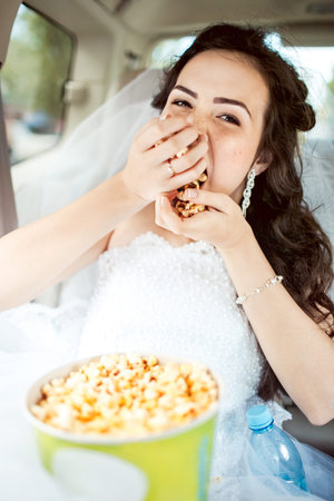 Portrait of beautiful young bride sitting in car straight, greedily eats popcorn before movie, hairstyle and bright makeup. Strong expression, shallow depth of field, focus on eyes and a bit on mouth.の写真素材