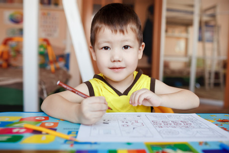 A three year boy in the yellow T-shirt with hood sits at the table, drawing and writing letters with color pencils, looking straight at the camera, interested gaze.の写真素材