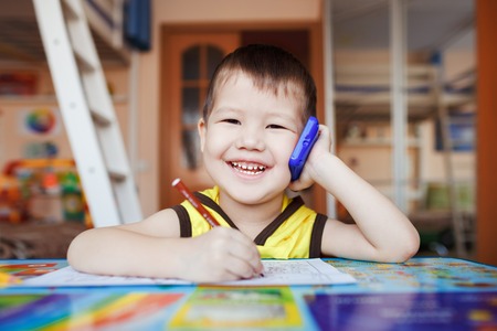 Happy little boy talking on smartphone at home while writing letters, three years old.の写真素材