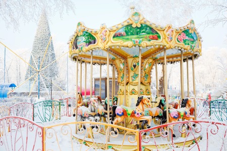 Merry-go-round with traditional horses covered with snow. Behind the carousel (roundabout) big Christmas tree. City park during the winter frosty day.の写真素材