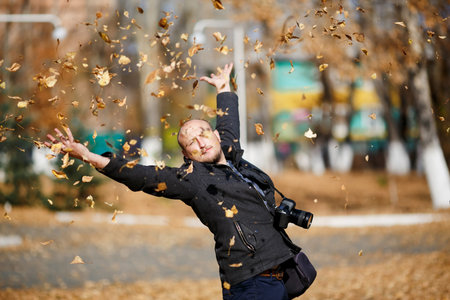 Portrait of a happy male photographer with professional digital camera on his neck, he throws tons of leaves falling in the air in autumn park, enjoying the sunshine weather, glad and satisfied.の写真素材