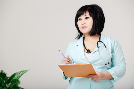 Curvy female nurse stands still writes the results by pen on paper, in a medical lab coat with a stethoscope around her neck. Asian appearance, dark short hair. Small vegetation in the foreground.の写真素材