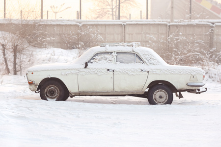 Abandoned car covered with snow in winter, old broken white, at sunset, warm tones, side view. Rusting, recycling, metal processing, write-off of cars.の写真素材