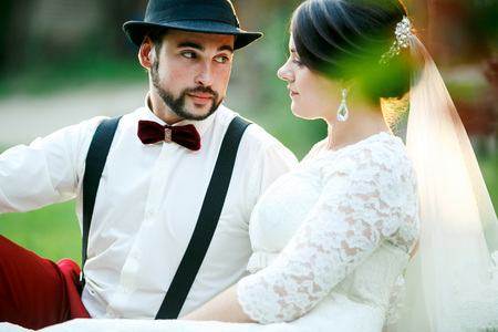 Stylish bride and groom sit on grass in rays of setting sunlight. Man in hat with bow tie and suspenders, red pants languidly looks at a woman with professional hair styling. A couple of newlyweds.の写真素材