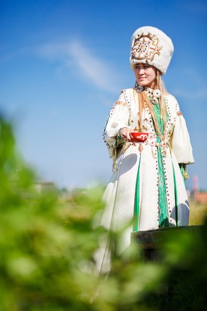 Pretty girl in national dress with red bowl in her hands outdoors on sunny dayの写真素材
