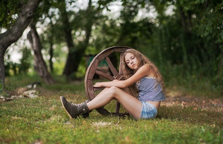 Summer day in the village. A girl sitting next to a wooden wheel.の写真素材