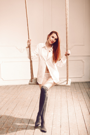Cute redhead woman with long hair sitting on swing in man's shirt long socks and sexy white lingerieの写真素材