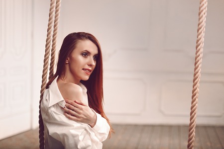 Cute young redhead woman with long hair sitting on swing in man's shirt long socks and sexy white lingerieの写真素材