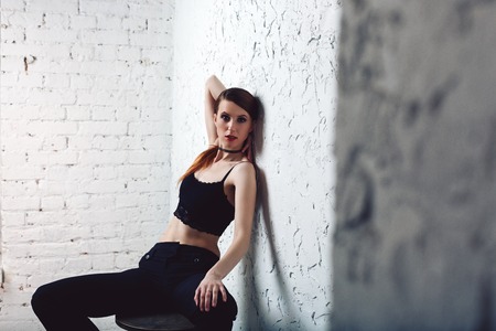 Attractive redhead model woman sitting in sexual pose on bar chair on white wall in sexy lace top and black jeansの写真素材