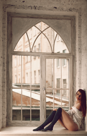 Sexy young redhead woman with long hair sitting on windowsill in man's shirt and socksの写真素材